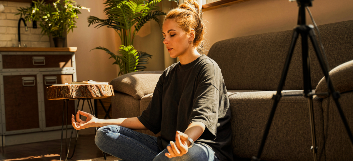 A person sits cross-legged on a couch in a sunlit room, meditating with relaxed hands and houseplants in the background.
