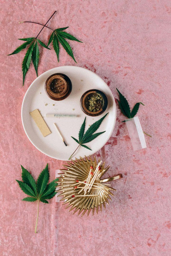A white tray holds cannabis leaves, a small container of ground flower, rolling papers, and smoking accessories on a pink textured surface.