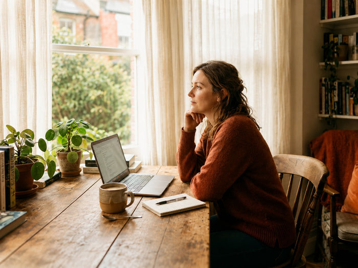 A woman in a rust-colored sweater sits at a wooden table with a laptop, notebook, coffee mug, and potted plants, gazing thoughtfully out a sunlit window in a cozy home workspace.