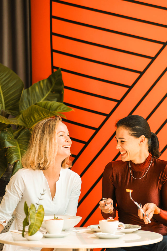 Two friends sit together chatting and smiling in front of a bright orange wall.