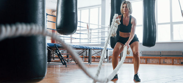 A woman in black activewear uses battle ropes in a bright gym with punching bags and a boxing ring in the background.