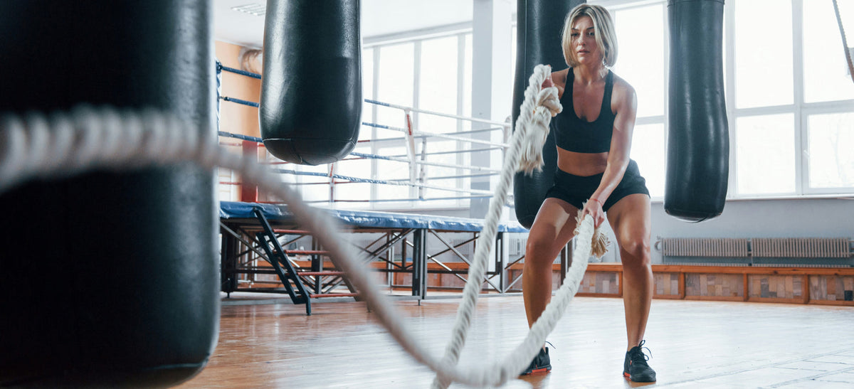 A woman in black activewear uses battle ropes in a bright gym with punching bags and a boxing ring in the background.