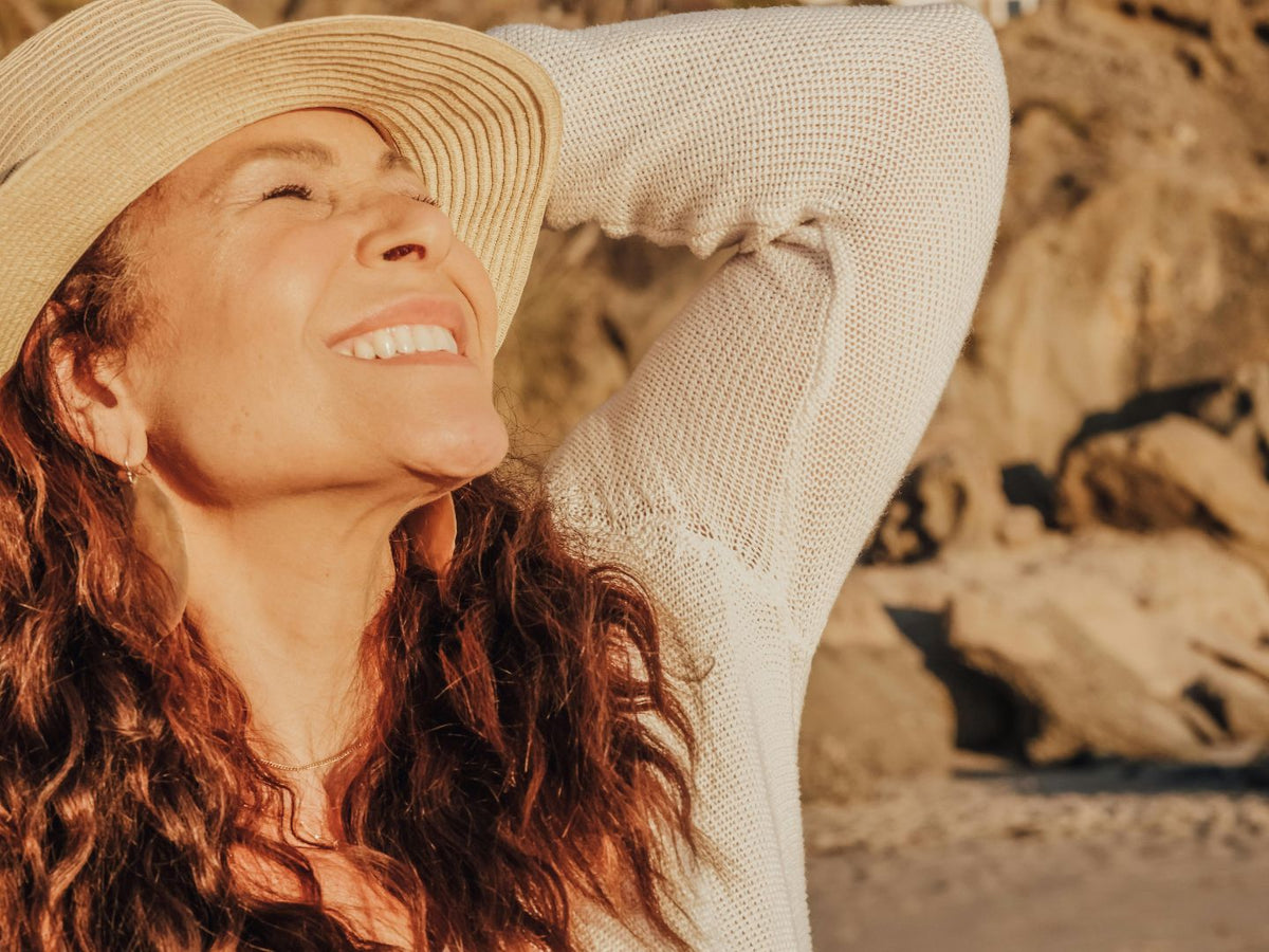 A smiling woman in a sunhat enjoys warm sunlight outdoors near a rocky beach.