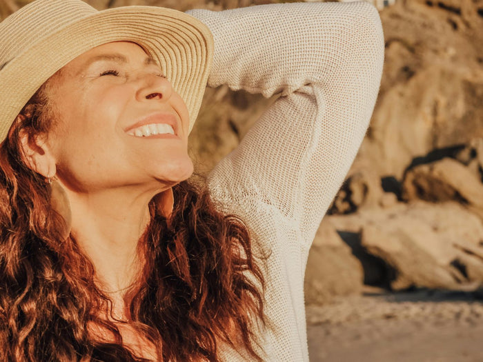 A smiling woman in a sunhat enjoys warm sunlight outdoors near a rocky beach.