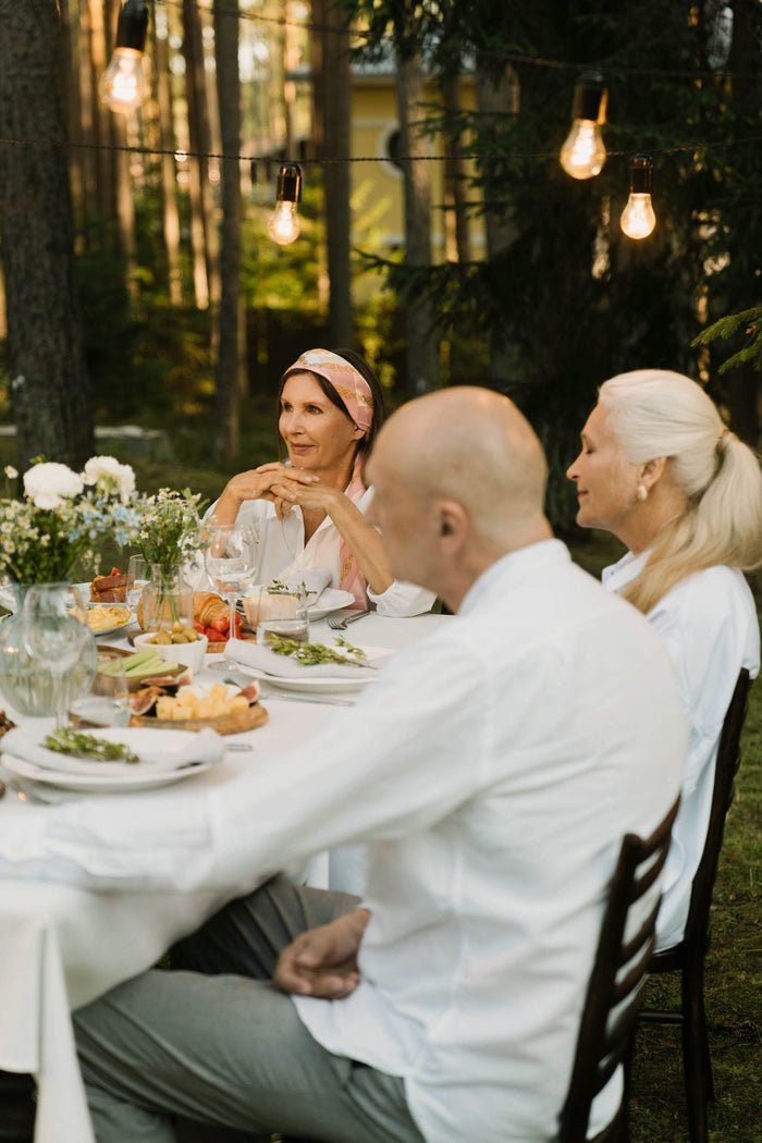 A group of adults gathers around an outdoor table set with food, flowers, and glassware in a relaxed garden setting.