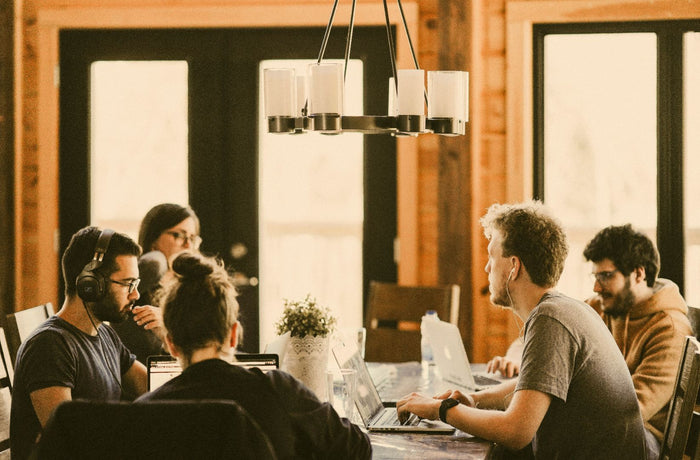 A group of people work together at laptops around a table in a warm, sunlit office space.