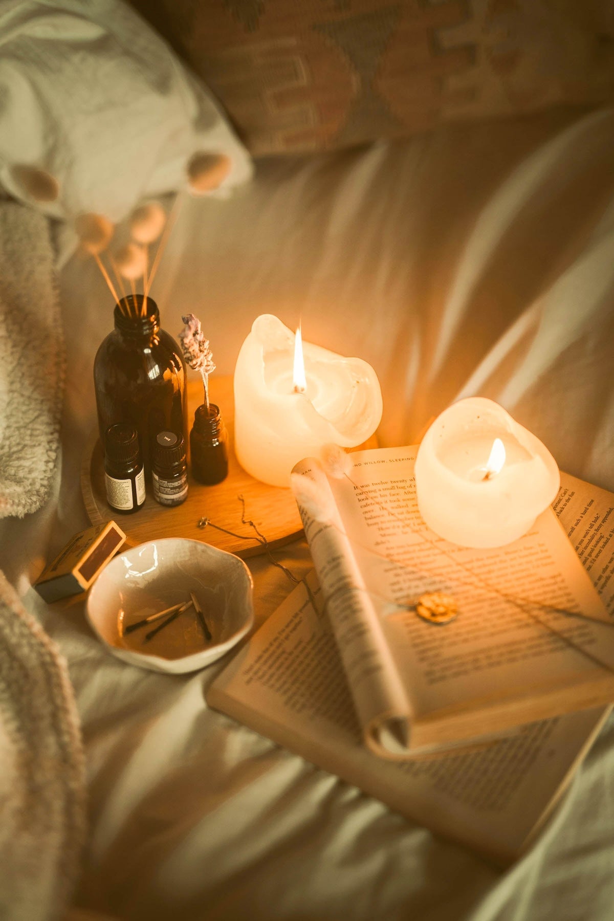 Lit candles, amber bottles, a dried flower, and an open book sit on a wooden tray in warm, cozy light.
