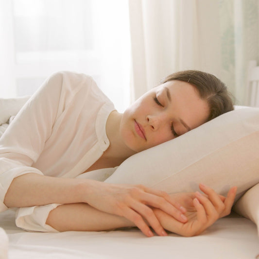 Woman sleeps peacefully on her side in a bright white bedroom, resting on a pillow under soft bedding with warm morning light.