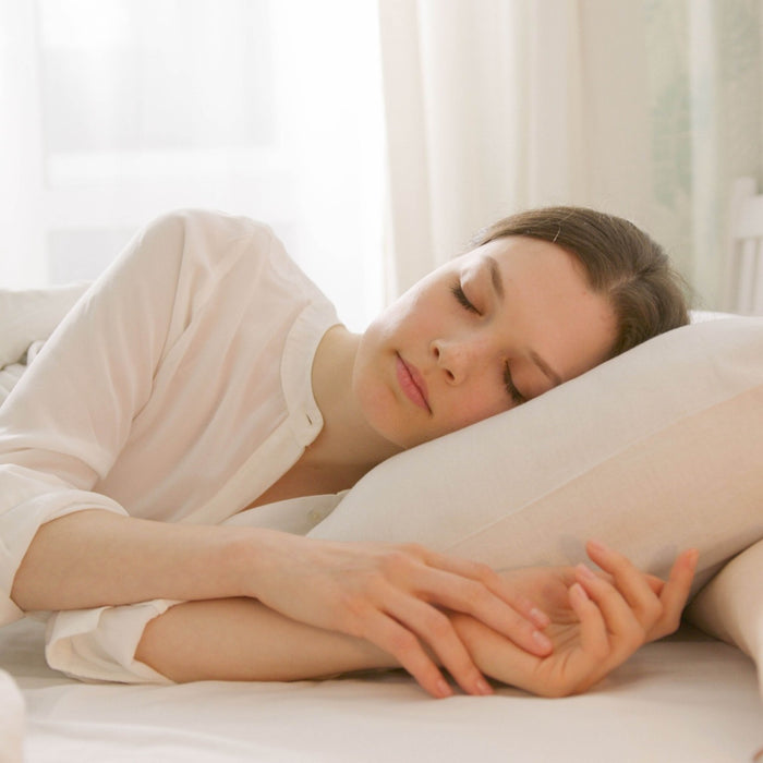 Woman sleeps peacefully on her side in a bright white bedroom, resting on a pillow under soft bedding with warm morning light.