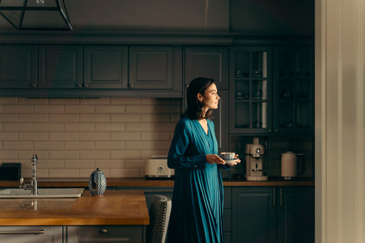 Woman in a long teal dress holding a teacup and saucer in a dim wood-and-tile kitchen