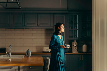 Woman in a long teal dress holding a teacup and saucer in a dim wood-and-tile kitchen