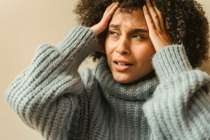 A person in a chunky gray sweater holds their head with both hands, looking stressed or overwhelmed in warm natural light.