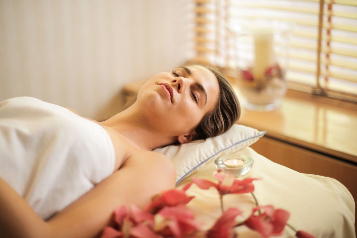 A woman relaxes with her eyes closed during a spa treatment, lying under a white towel with soft candles, flowers, and warm sunlight nearby.
