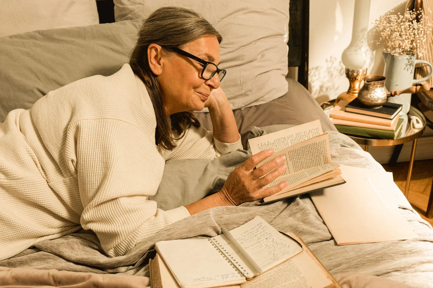 Woman relaxing in bed while reading a book in warm evening light, capturing a calm, cozy routine that helps her ease into rest at her own pace