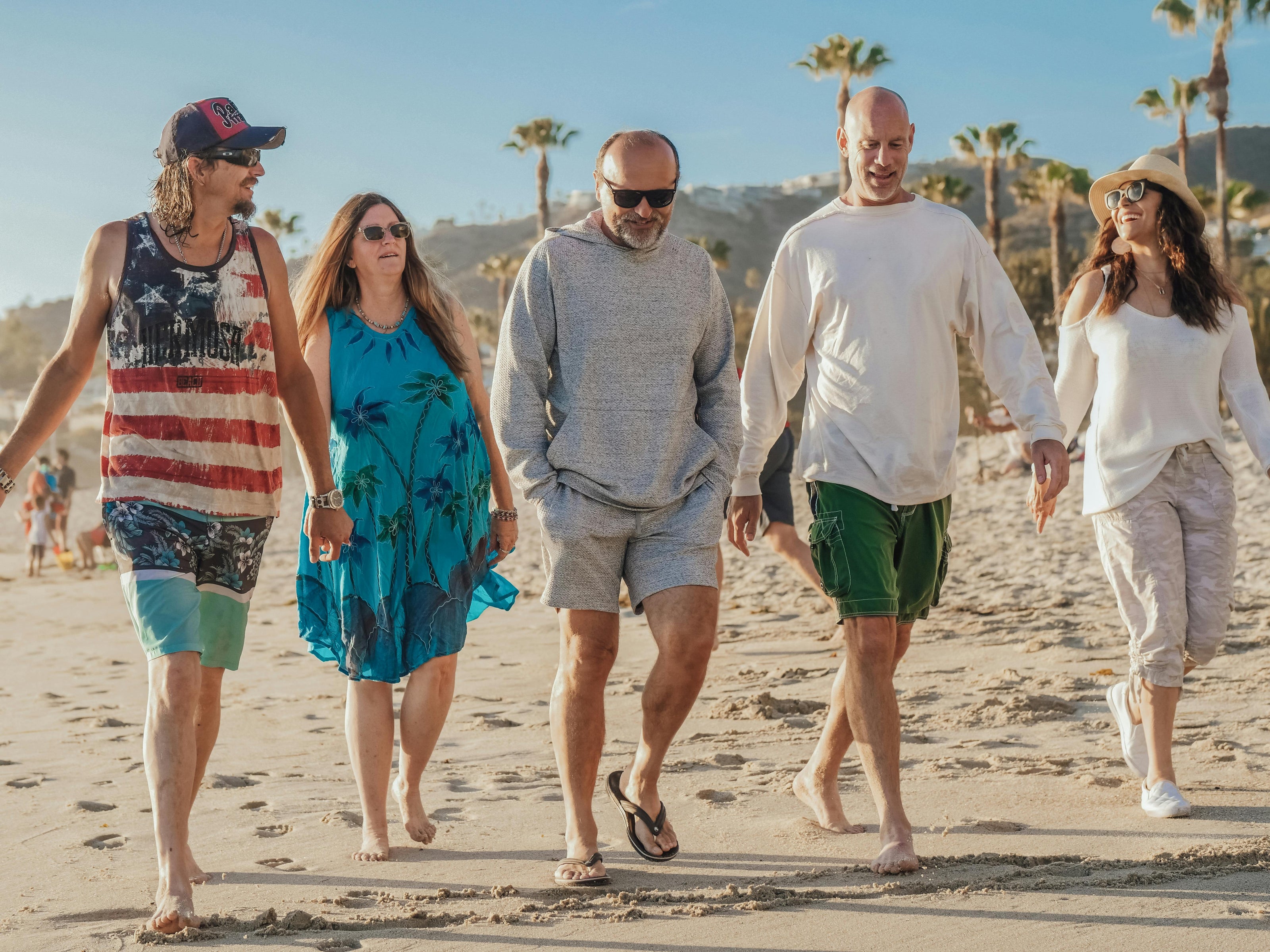 Five adults walking together along a sandy beach in daylight, with palm trees and hills in the background, dressed in casual warm-weather clothing