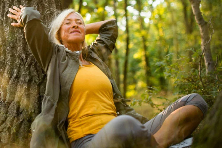 Woman resting against a tree in a sunlit forest, eyes closed and shoulders relaxed, taking a quiet moment to unwind and recharge outdoors