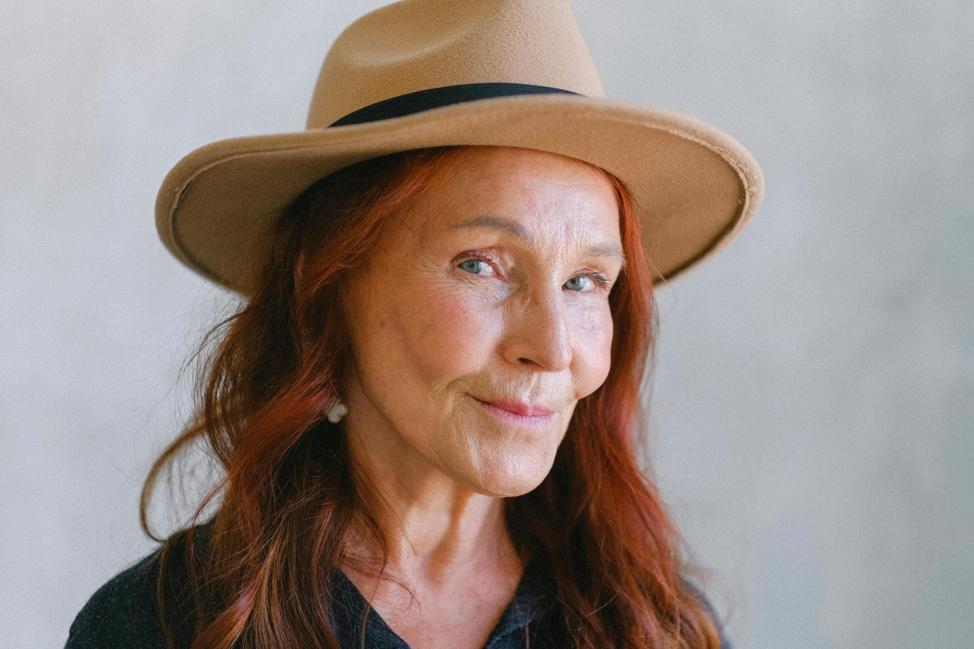 Close-up portrait of a woman with red hair wearing a wide-brim hat, looking at the camera with a soft smile against a neutral background